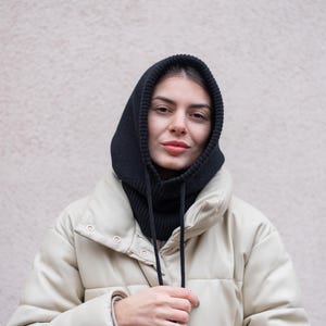 Puede incluir: Una mujer con un cuello de lana negro con capucha y una chaqueta acolchada blanca. El cuello tiene un cord&oacute;n.
