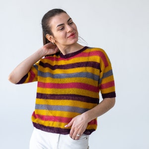 May include: A woman wearing a striped sweater with yellow, pink, purple, and gray stripes. She is wearing white pants and has her hand in her pocket.
