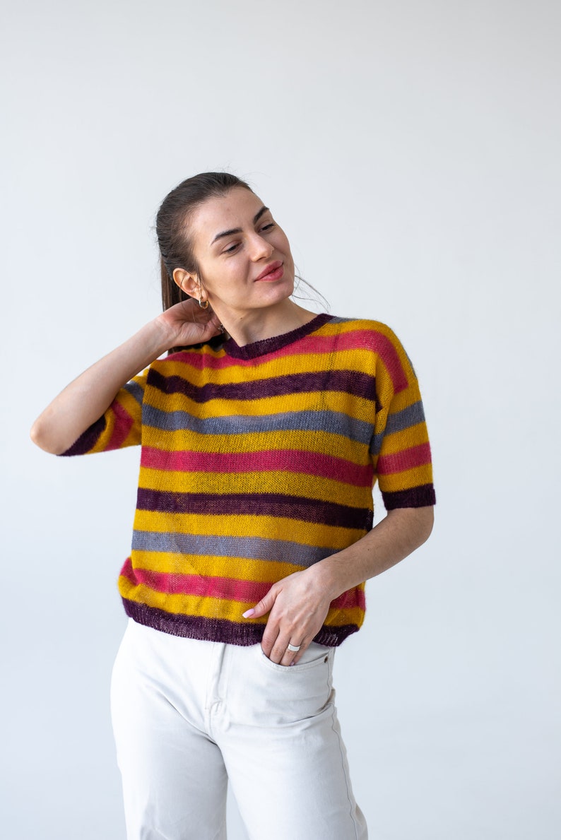 May include: A woman wearing a striped sweater with yellow, pink, purple, and gray stripes. She is wearing white pants and has her hand in her pocket.