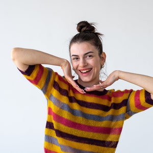 May include: A woman with a bun in her hair is wearing a yellow, pink, purple, and gray striped sweater. She is smiling and has her hands on her cheeks.