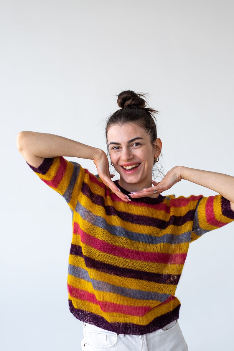 May include: A woman with a bun in her hair is wearing a yellow, pink, purple, and gray striped sweater. She is smiling and has her hands on her cheeks.