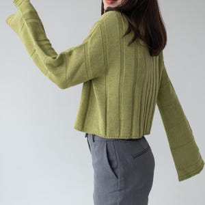 May include: A cropped, light green sweater with flared sleeves and vertical ribbed detailing. The model is also wearing gray trousers. The sweater's color is a muted, earthy tone, and the sleeves have a bell-like shape.