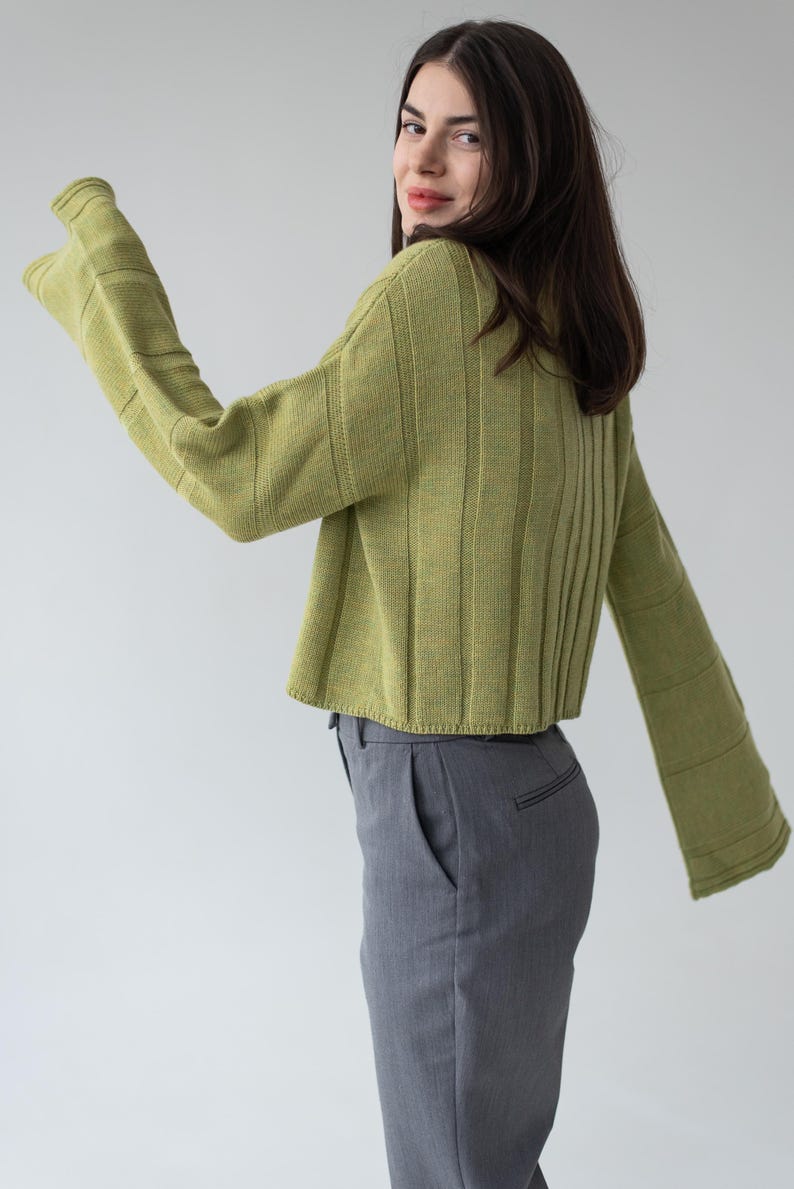 May include: A cropped, light green sweater with flared sleeves and vertical ribbed detailing. The model is also wearing gray trousers. The sweater's color is a muted, earthy tone, and the sleeves have a bell-like shape.