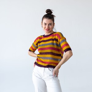 May include: A woman with her hair pulled back in a bun is wearing a striped sweater with yellow, pink, and purple stripes. She is also wearing white pants.