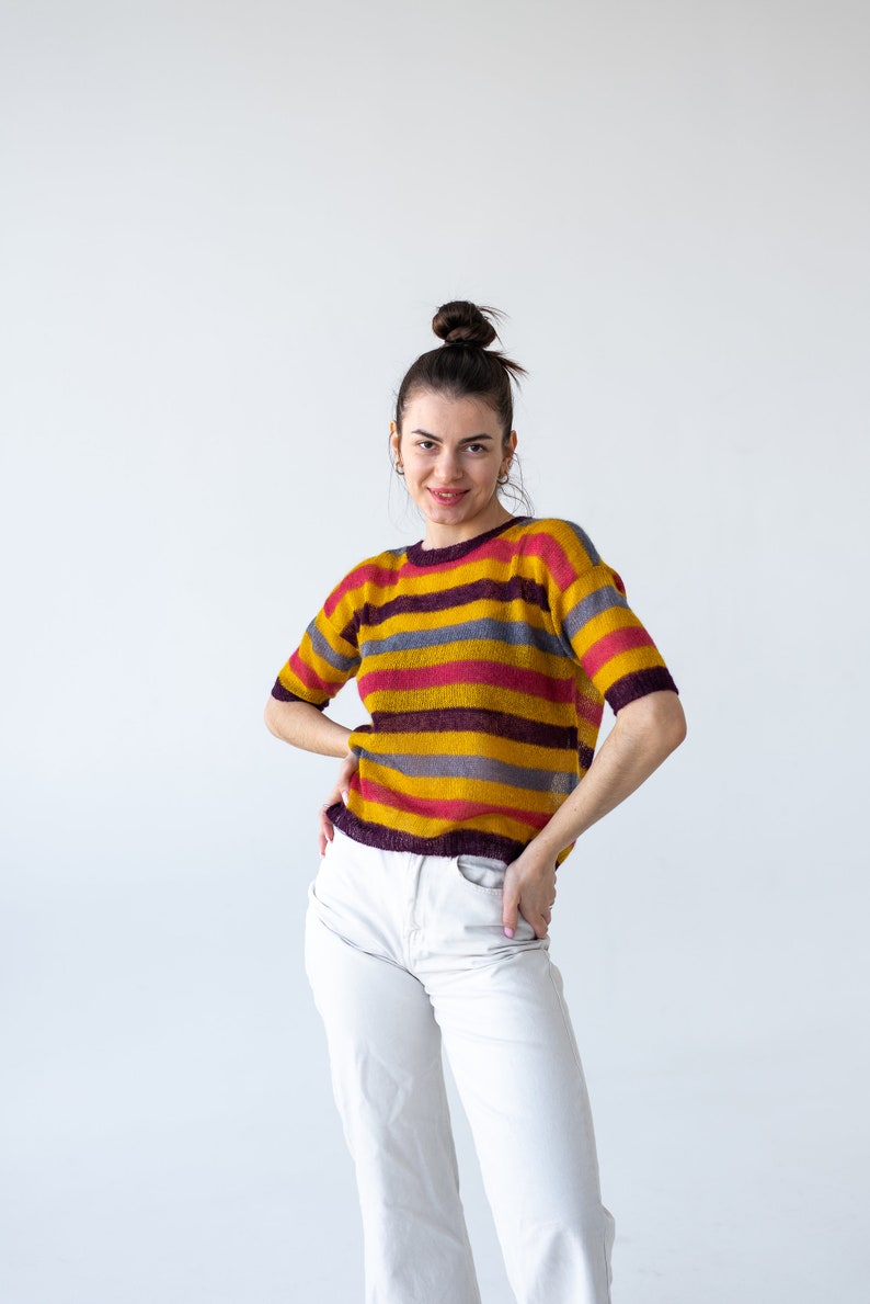 May include: A woman with her hair pulled back in a bun is wearing a striped sweater with yellow, pink, and purple stripes. She is also wearing white pants.