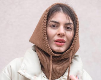May include: A woman wearing a brown knitted hooded scarf over a cream colored puffer jacket.