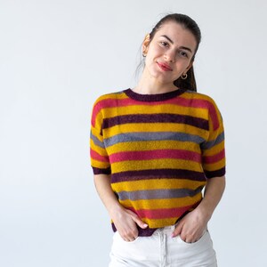 May include: A woman wearing a yellow, pink, and gray striped sweater with white pants. She is standing in front of a white background.