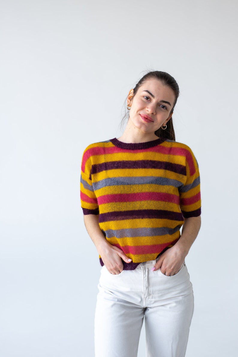 May include: A woman wearing a yellow, pink, and gray striped sweater with white pants. She is standing in front of a white background.