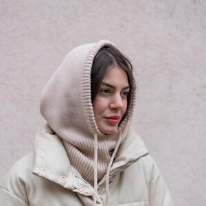 Puede incluir: Una mujer con una chaqueta acolchada de color crema con una bufanda de capucha de punto a juego. La chaqueta tiene una cremallera y un cierre de presi&oacute;n.