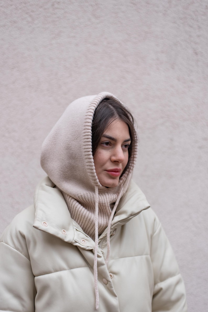 Puede incluir: Una mujer con una chaqueta acolchada de color crema con una bufanda de capucha de punto a juego. La chaqueta tiene una cremallera y un cierre de presi&oacute;n.