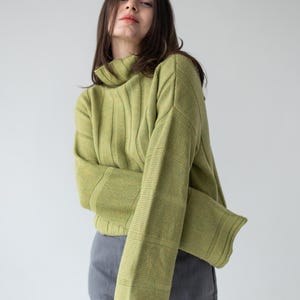 May include: A light green ribbed knit sweater with a high neck and long sleeves. The sweater is paired with gray pants. The model is facing the camera.