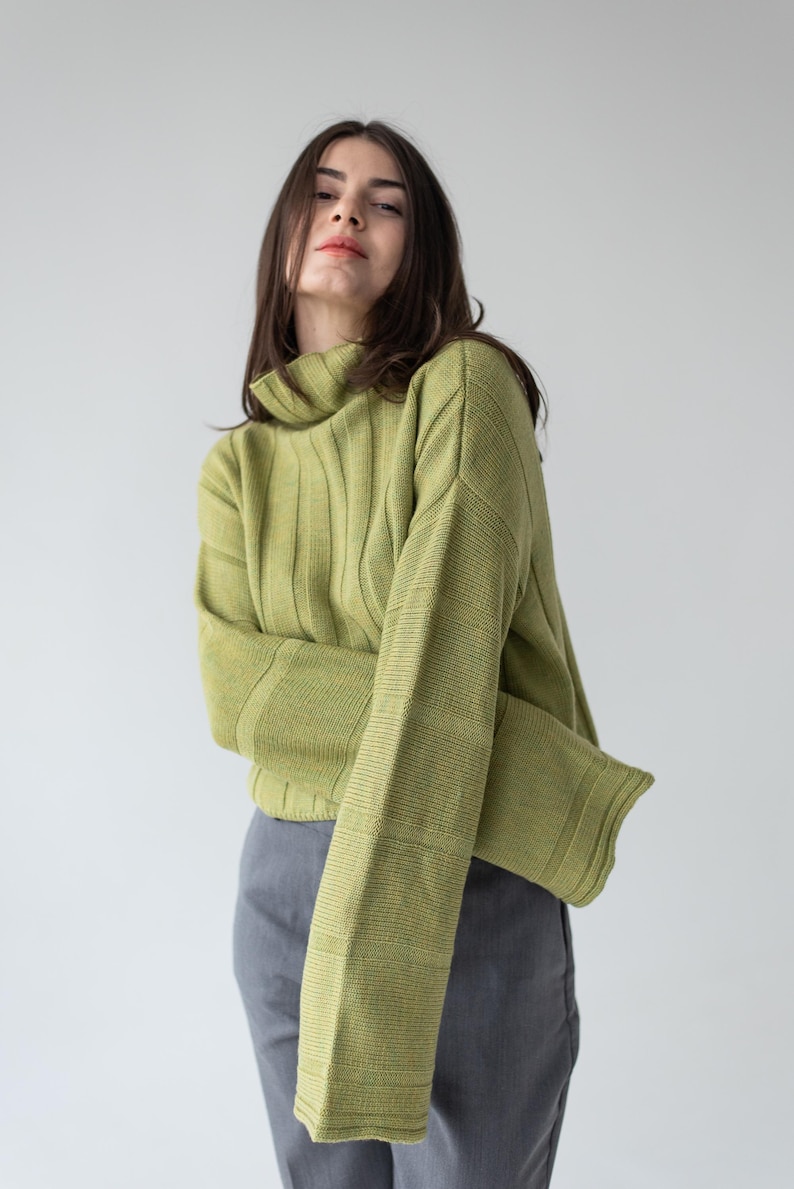 May include: A light green ribbed knit sweater with a high neck and long sleeves. The sweater is paired with gray pants. The model is facing the camera.