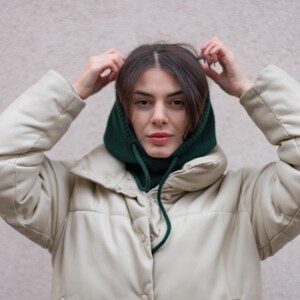 May include: A woman wearing a white puffer jacket with a green knit neck warmer. She is adjusting the neck warmer with her hands.