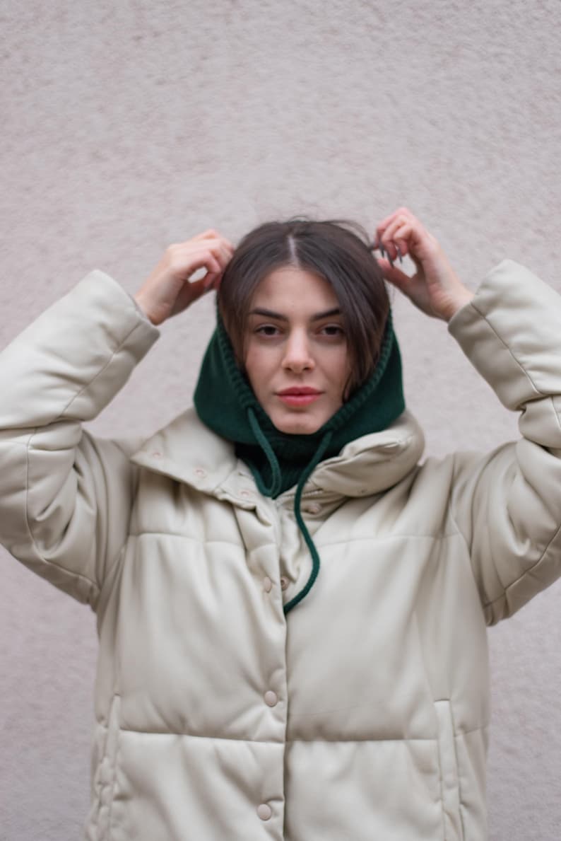 May include: A woman wearing a white puffer jacket with a green knit neck warmer. She is adjusting the neck warmer with her hands.