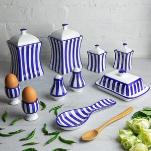 May include: A set of blue and white striped ceramic kitchenware, including canisters, salt and pepper shakers, a butter dish, and a spoon rest. The set also includes two egg cups and a wooden spoon.