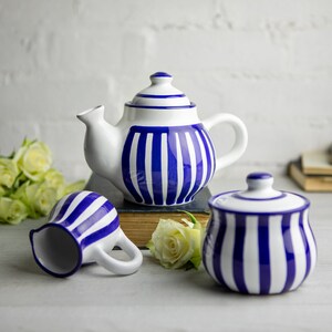 Può includere: Un set di tre articoli da cucina in ceramica a strisce blu e bianche: una teiera, un creamer e una zuccheriera. La teiera ha un manico e un beccuccio. Il creamer e la zuccheriera hanno i coperchi.