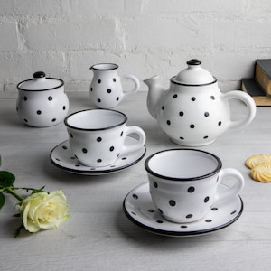 May include: A white teapot, creamer, sugar bowl, and two teacups and saucers with black polka dots and black trim. The tea set is on a white wood surface.