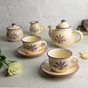 May include: A lavender-themed tea set with a teapot, creamer, sugar bowl, and two teacups and saucers. The set is decorated with purple lavender flowers and a light pink stripe.