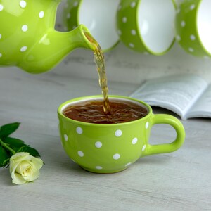 May include: A green teacup with white polka dots is being filled with tea from a green teapot with white polka dots. The teacup and teapot are on a white surface.