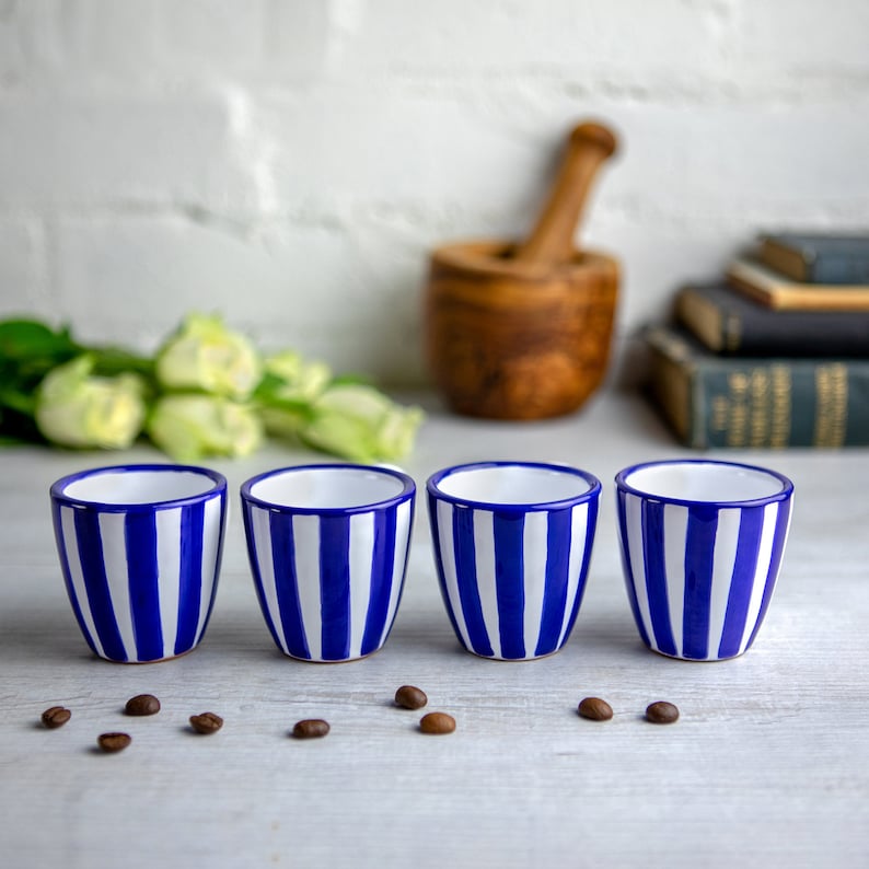 May include: Four white ceramic cups with blue stripes. The cups are arranged in a row on a white surface.