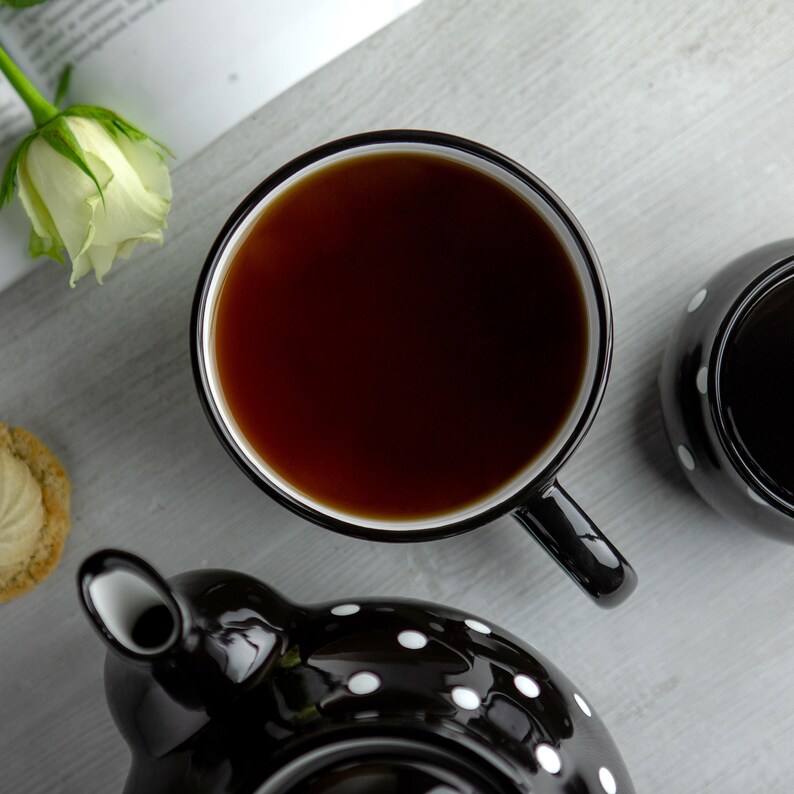 K&ouml;nnte beinhalten: Eine Teekanne und eine passende Teetasse mit schwarzen und wei&szlig;en Punkten. Die Teetasse ist mit schwarzem Tee gef&uuml;llt.