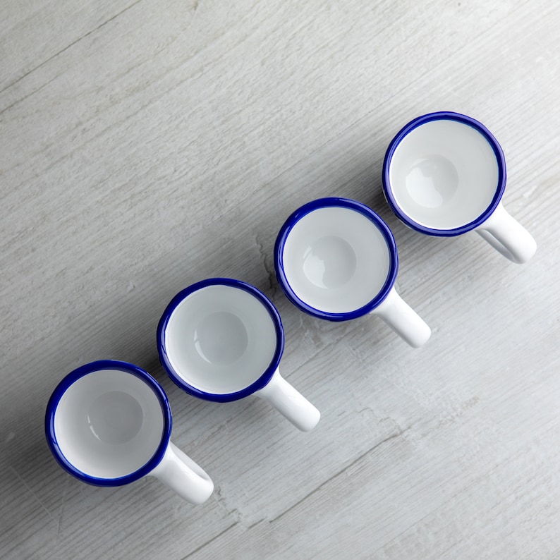 May include: Four white enamel mugs with blue rims. The mugs have a rounded shape and a small handle on the side. The mugs are arranged in a row on a light wood surface.