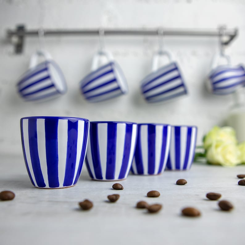 May include: Four blue and white striped ceramic mugs with a white background. The mugs are arranged in a row on a white surface.