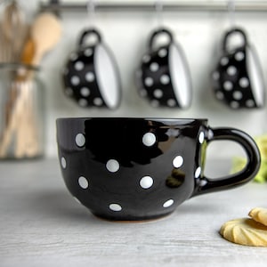 K&ouml;nnte beinhalten: Eine schwarze Keramiktasse mit wei&szlig;en Punkten. Die Tasse hat eine abgerundete Form und einen Henkel. Sie steht auf einer wei&szlig;en Oberfl&auml;che.