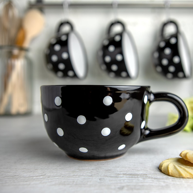 K&ouml;nnte beinhalten: Eine schwarze Keramiktasse mit wei&szlig;en Punkten. Die Tasse hat eine abgerundete Form und einen Henkel. Sie steht auf einer wei&szlig;en Oberfl&auml;che.