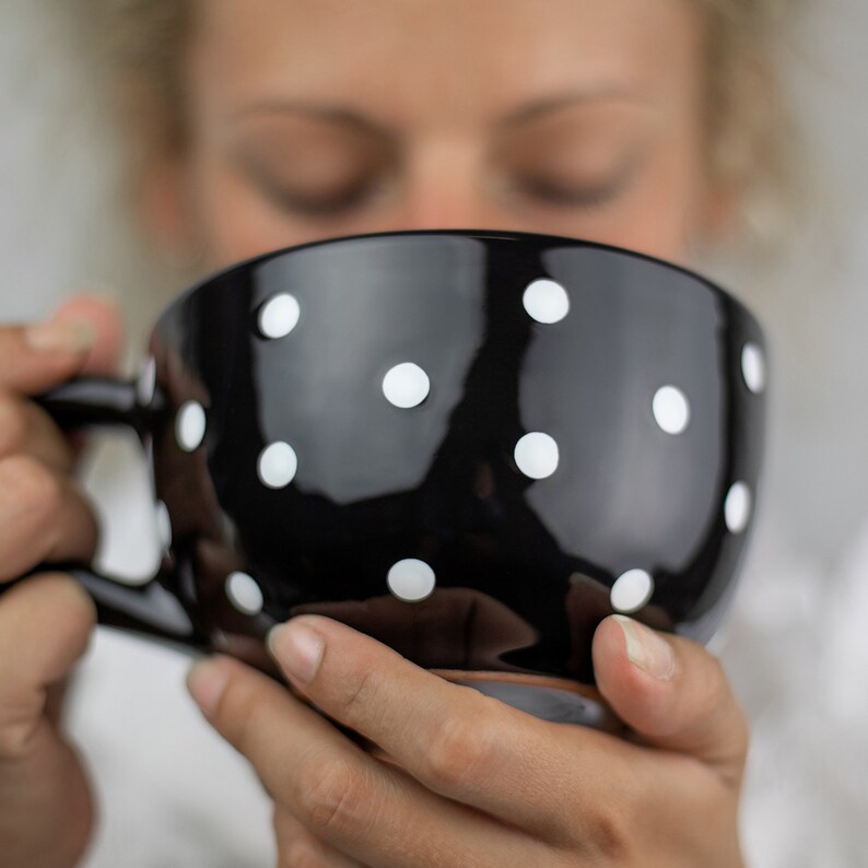 K&ouml;nnte beinhalten: Eine schwarze Keramiktasse mit wei&szlig;en Punkten. Die Tasse wird von einer Person gehalten.