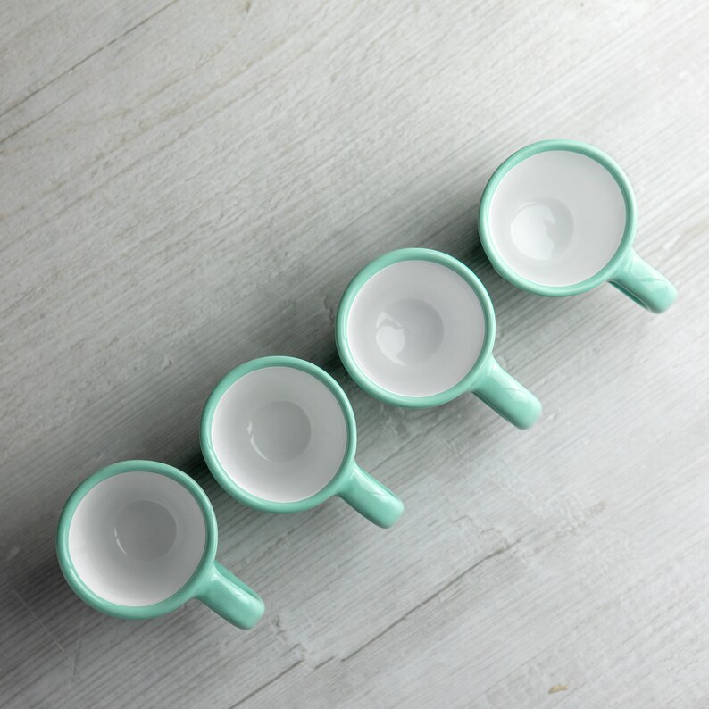 May include: Five light blue ceramic mugs with white interiors lined up in a row on a light wood surface.
