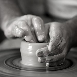 May include: A person's hands shaping a clay pot on a pottery wheel. The clay is smooth and wet, and the potter's hands are covered in clay.
