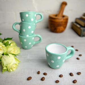 May include: Four mint green espresso cups with white polka dots stacked on a white surface with coffee beans scattered around.