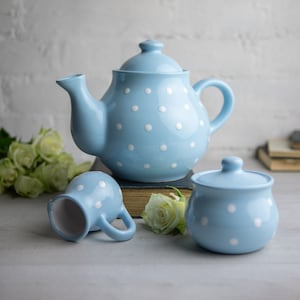 Può includere: Una teiera, un bricco per la panna e una zuccheriera blu con pois bianchi. La teiera è appoggiata su un libro. Il bricco per la panna e la zuccheriera sono su una superficie bianca.