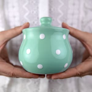 Peut inclure: Un pot à sucre en céramique vert menthe avec un couvercle blanc et des pois blancs.