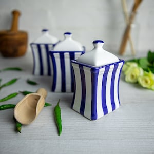 May include: Three white ceramic canisters with blue stripes. The canisters are arranged in a row on a white surface. The canisters have lids and are ideal for storing spices, sugar, or other kitchen essentials.