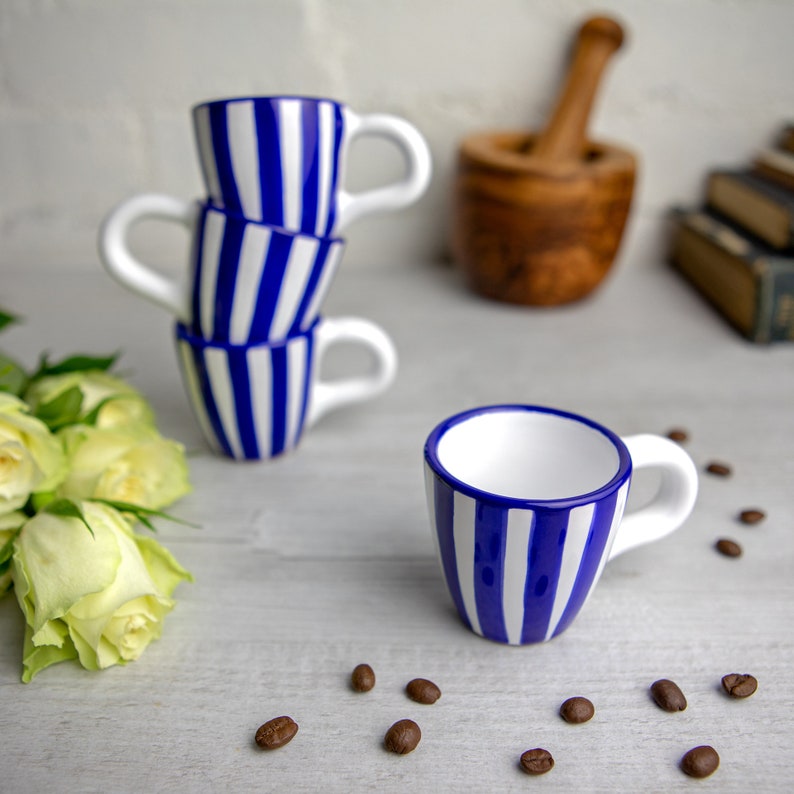 May include: Four blue and white striped ceramic espresso cups stacked on top of each other. The top cup is tilted slightly to the side. A single espresso cup is sitting on a white surface with coffee beans scattered around it.