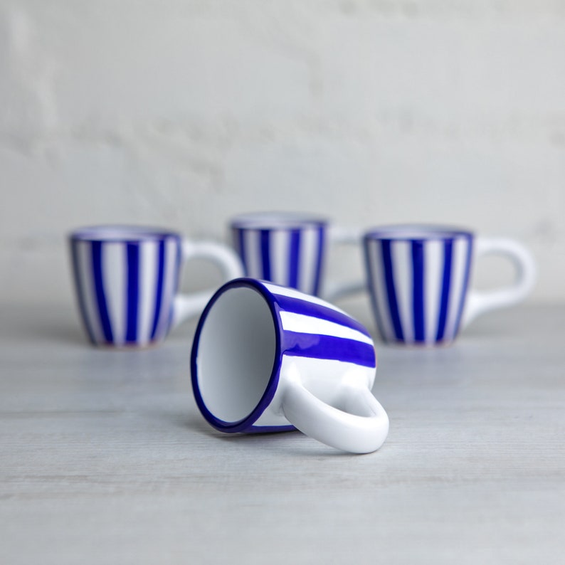 May include: Four white ceramic mugs with blue stripes. One mug is lying on its side, while the other three are standing upright.
