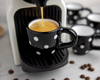 Op de afbeelding: Een witte espressomachine met een zwart-wit gepolka-dotte espressokopje gevuld met koffie. De koffie wordt uit de machine in het kopje gegoten.