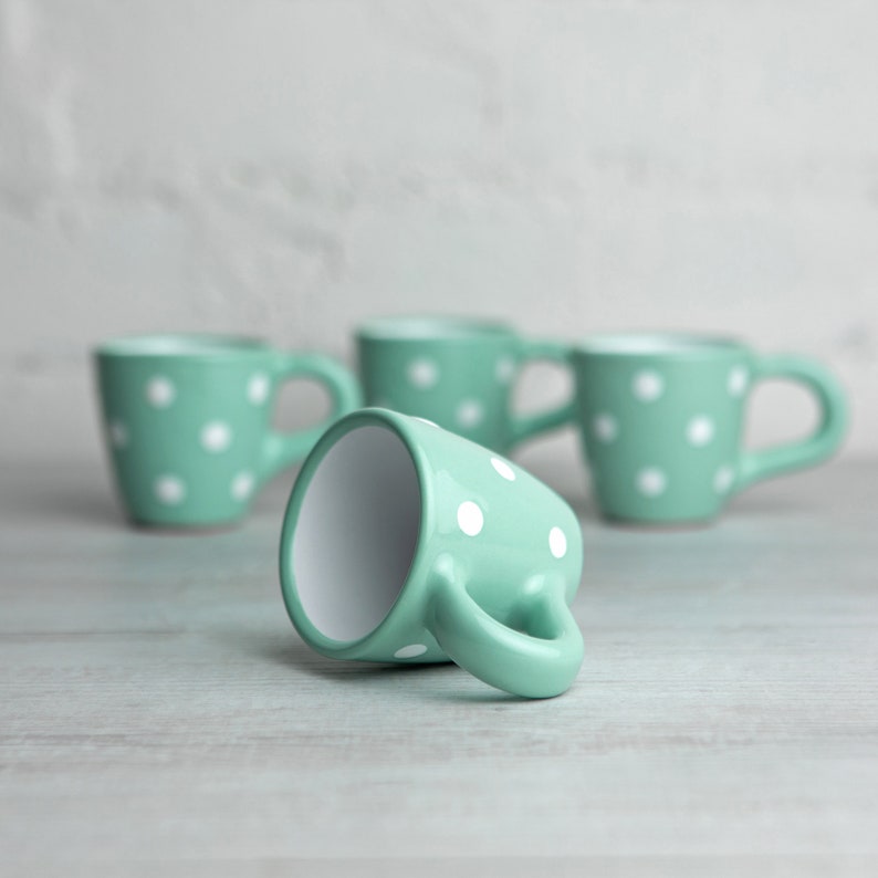 May include: Four mint green ceramic mugs with white polka dots. One mug is lying on its side, while the other three are standing upright.