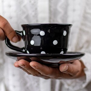 May include: A black teacup and saucer with white polka dots. The cup is held in a person's hand.