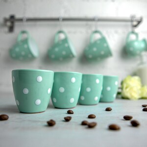 May include: Four mint green ceramic mugs with white polka dots, sitting on a white surface with coffee beans scattered around them.  The mugs are similar to four mugs hanging on a rack in the background.