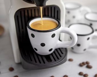 May include: A white espresso machine with a black drip tray pouring coffee into a white cup with black polka dots. The cup has a black rim and handle. There are several other white cups with black polka dots on the table.