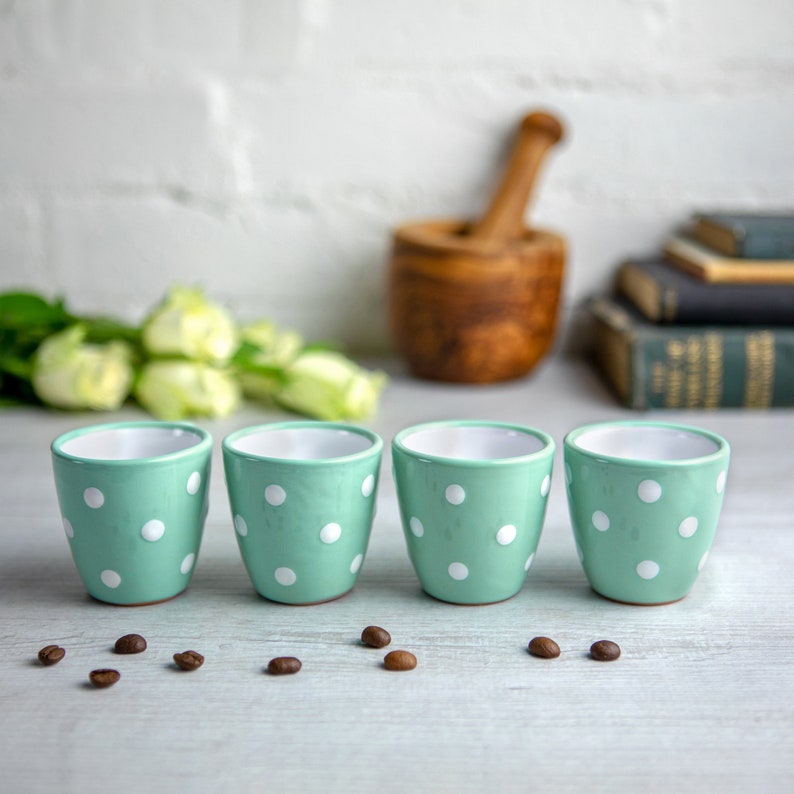 May include: Four mint green ceramic mugs with white polka dots. The mugs are lined up in a row on a white surface.