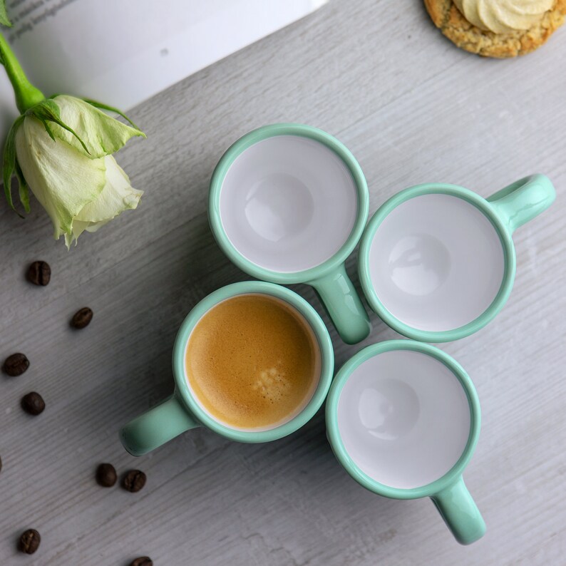 May include: Four light green ceramic espresso cups with white interiors. One cup is filled with coffee. The cups are arranged in a circle on a light gray surface.
