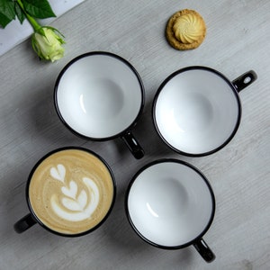 K&ouml;nnte beinhalten: Vier wei&szlig;e Keramikbecher mit schwarzen R&auml;ndern und Griffen. Drei Becher sind leer, und ein Becher hat ein Latte-Art-Design.