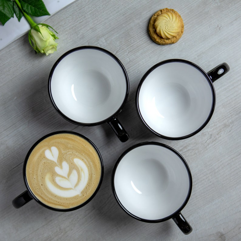 K&ouml;nnte beinhalten: Vier wei&szlig;e Keramikbecher mit schwarzen R&auml;ndern und Griffen. Drei Becher sind leer, und ein Becher hat ein Latte-Art-Design.