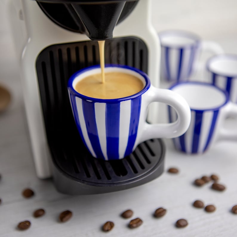 May include: A white and blue striped espresso cup with a handle is being filled with coffee from a white espresso machine. The cup is sitting on a black drip tray.