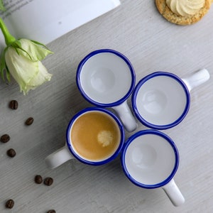 May include: Four white ceramic espresso cups with blue rims, three are empty and one is filled with coffee. The cups are arranged in a circle on a light gray surface.