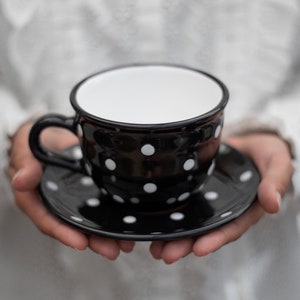 May include: A black teacup and saucer with white polka dots. The cup and saucer are held in a person's hands.